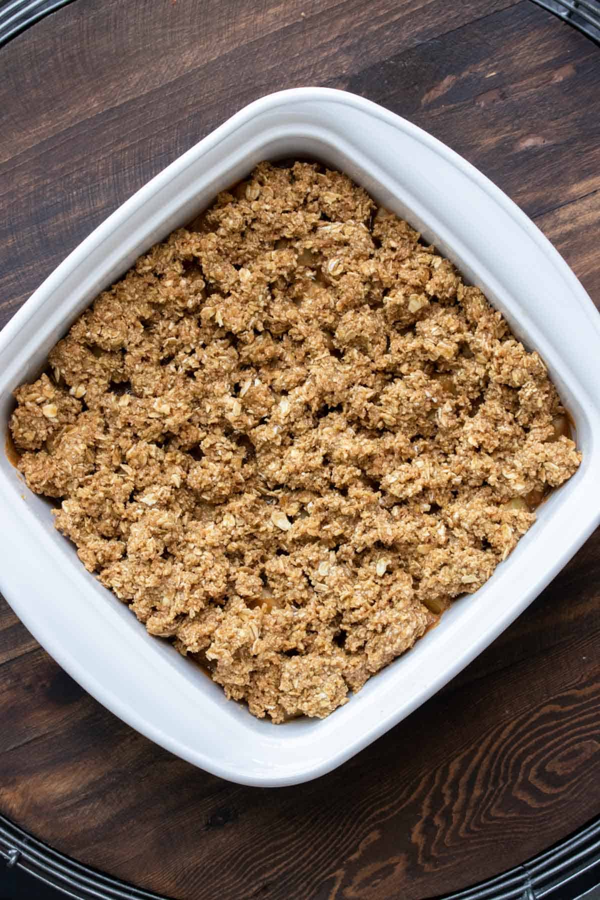 A white square dish with an unbaked apple crisp in it sitting on a wooden surface.