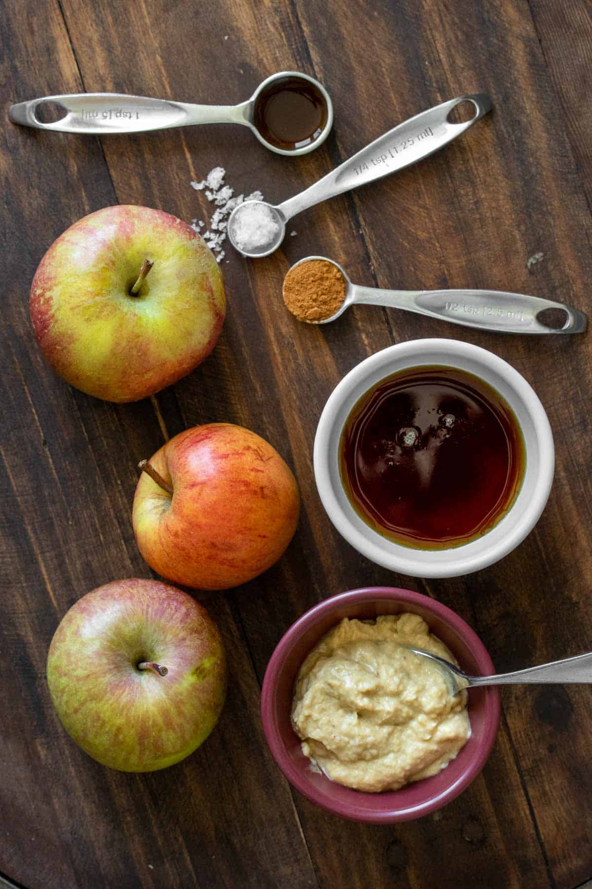 Top view of three apples next to bowls of cashew butter and maple syrup with spoons of seasonings.