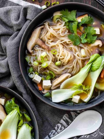 Asian style veggies and noodles in a broth in a black bowl with a white asian soup spoon next to it.