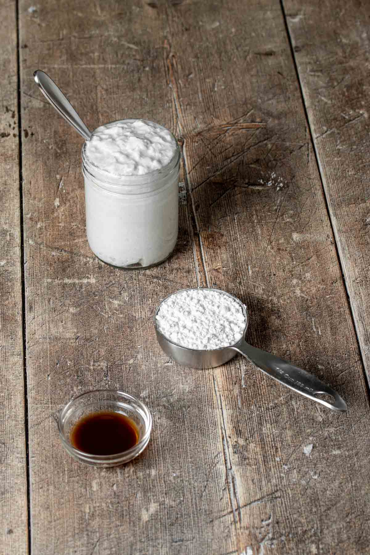 A wooden table with a jar of coconut cream, measuring cup of powdered sugar and glass bowl of vanilla extract.