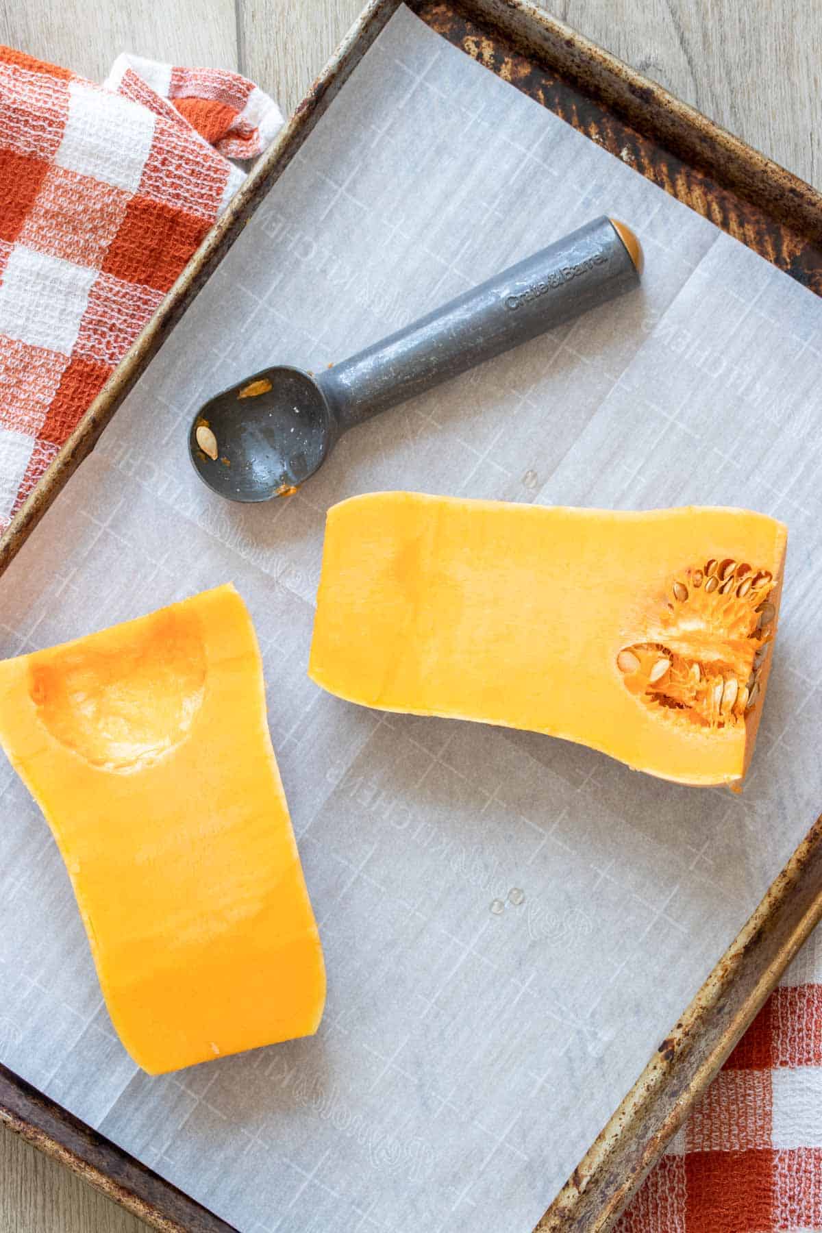 A parchment lined baking sheet with two halves of a butternut squash and an ice cream scooper.