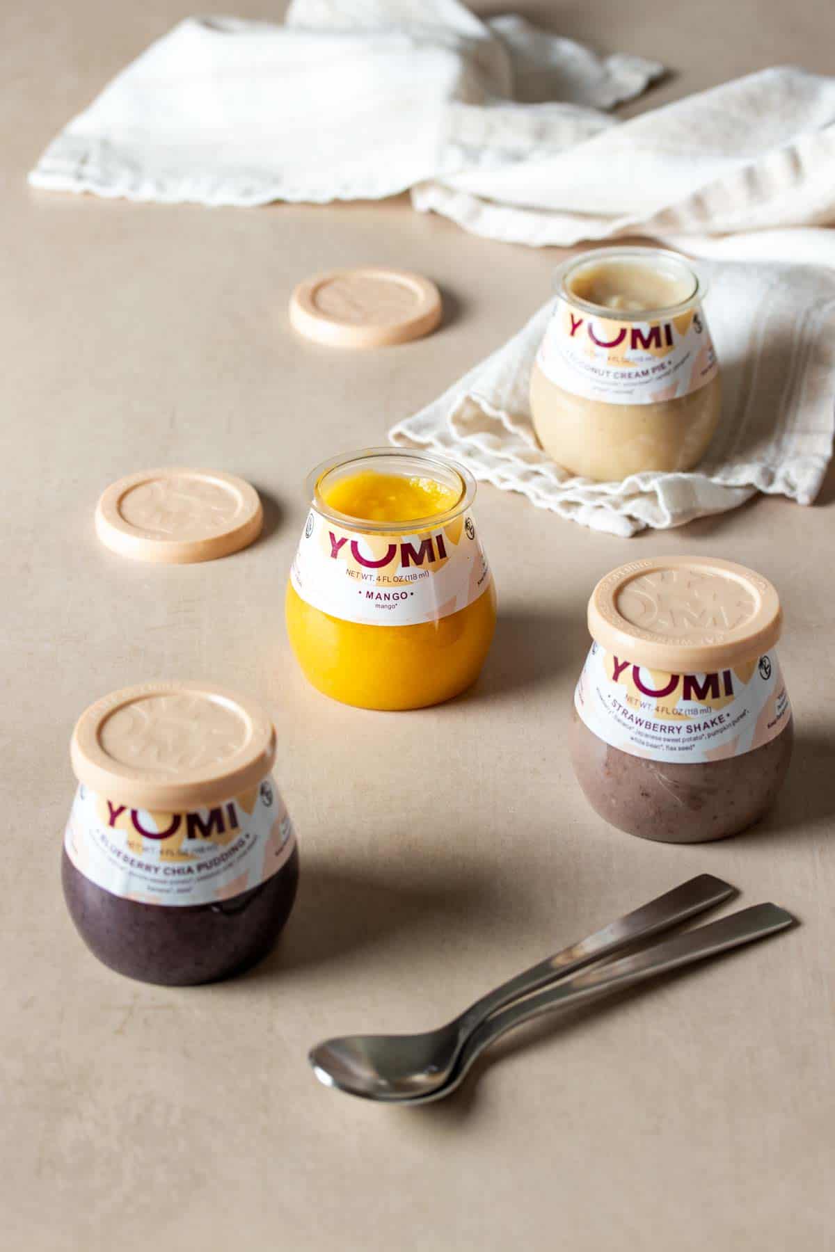 Four jars of baby food on a tan surface, two open with a set up spoons in front and a white towel in the back.