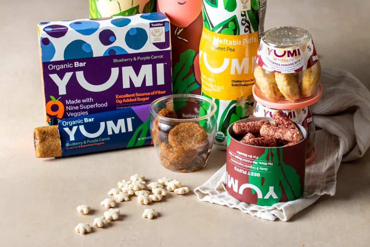 Colorful boxes, containers and jars of baby and toddler snacks, puffs and bars on a tan colored surface with a white towel.