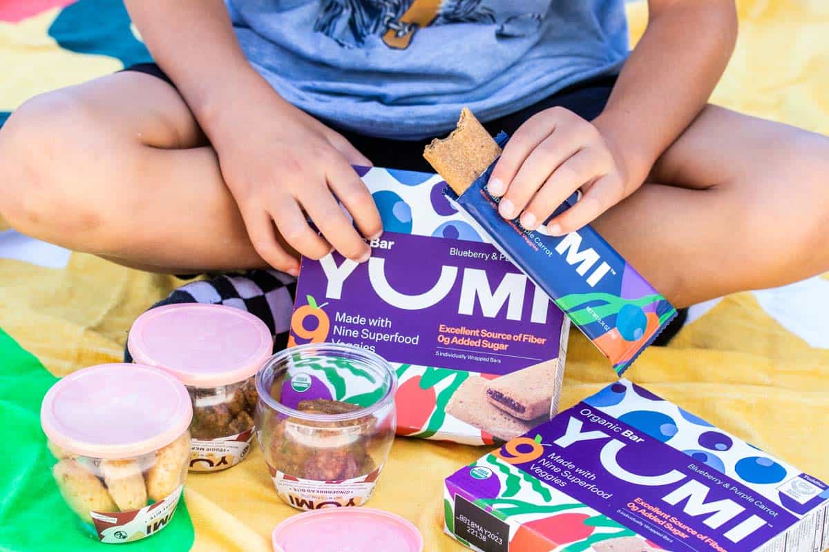 A boy sitting cross legged on a yellow towel with jars of snacks and bars in his lap and hands.