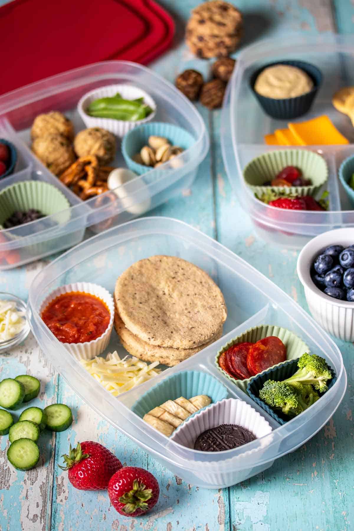 A light blue wooden surface with different fruit, veggies, grains and snacks in containers with sections.