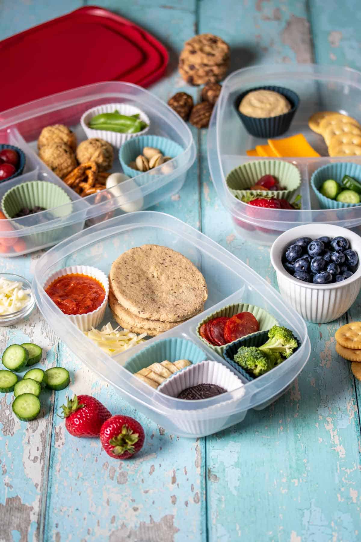 Fruit, veggies, grains, proteins and snacks inside containers with sections on a turquoise wooden surface.