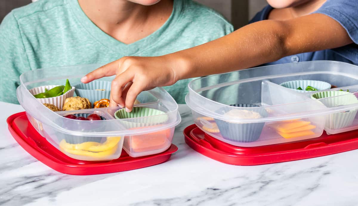 A boy taking something from a cupcake liner that is in a container with sections filled with fruit, veggies and snacks.