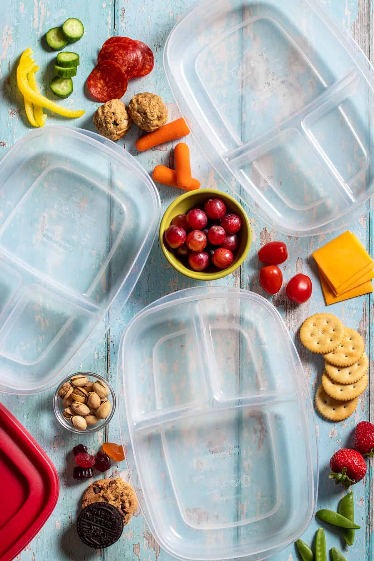 Top view of three empty plastic containers with sections surrounded by fruit, veggies, proteins, cheeses and snacks.