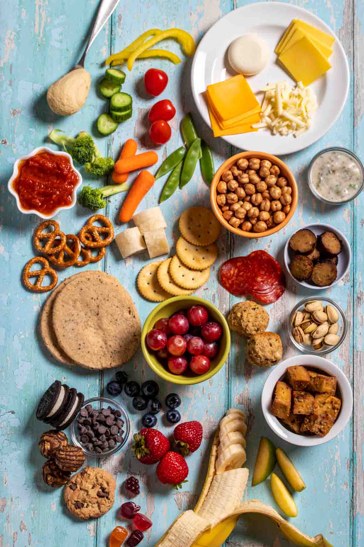 Top view of different kinds of fruit, veggies, grains, proteins, cheese and sauces lying on a turquoise surface.