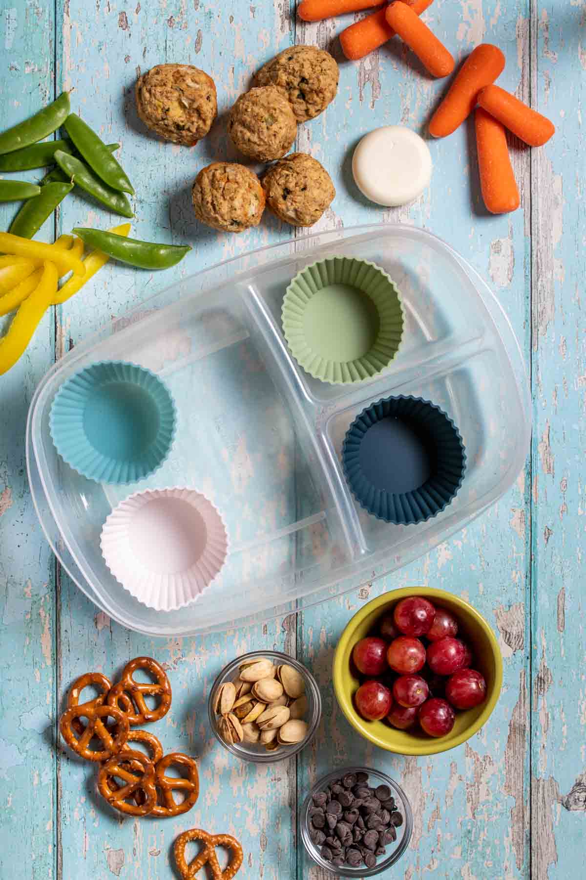 Top view of a sectioned off plastic container with cupcake liners inside to hold different foods surrounded by fruit, veggies and snacks.