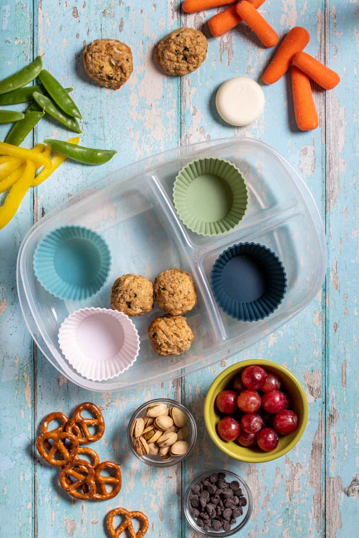 Top view of a plastic container with sections and cupcake liners and protein balls in the container surrounded by fruit, veggies and other snacks.