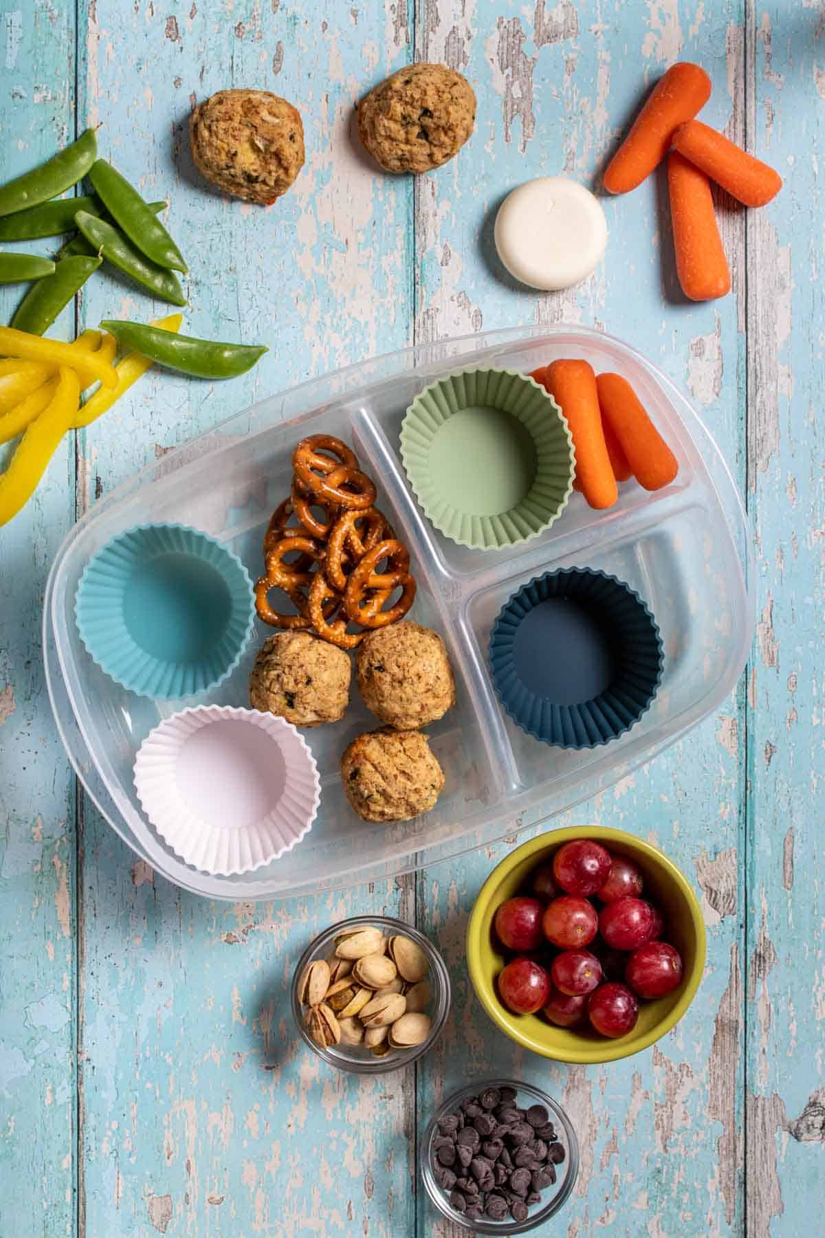 Top view of a container with sections with carrots, pretzels and protein balls with other fruit, veggies and snacks around it.