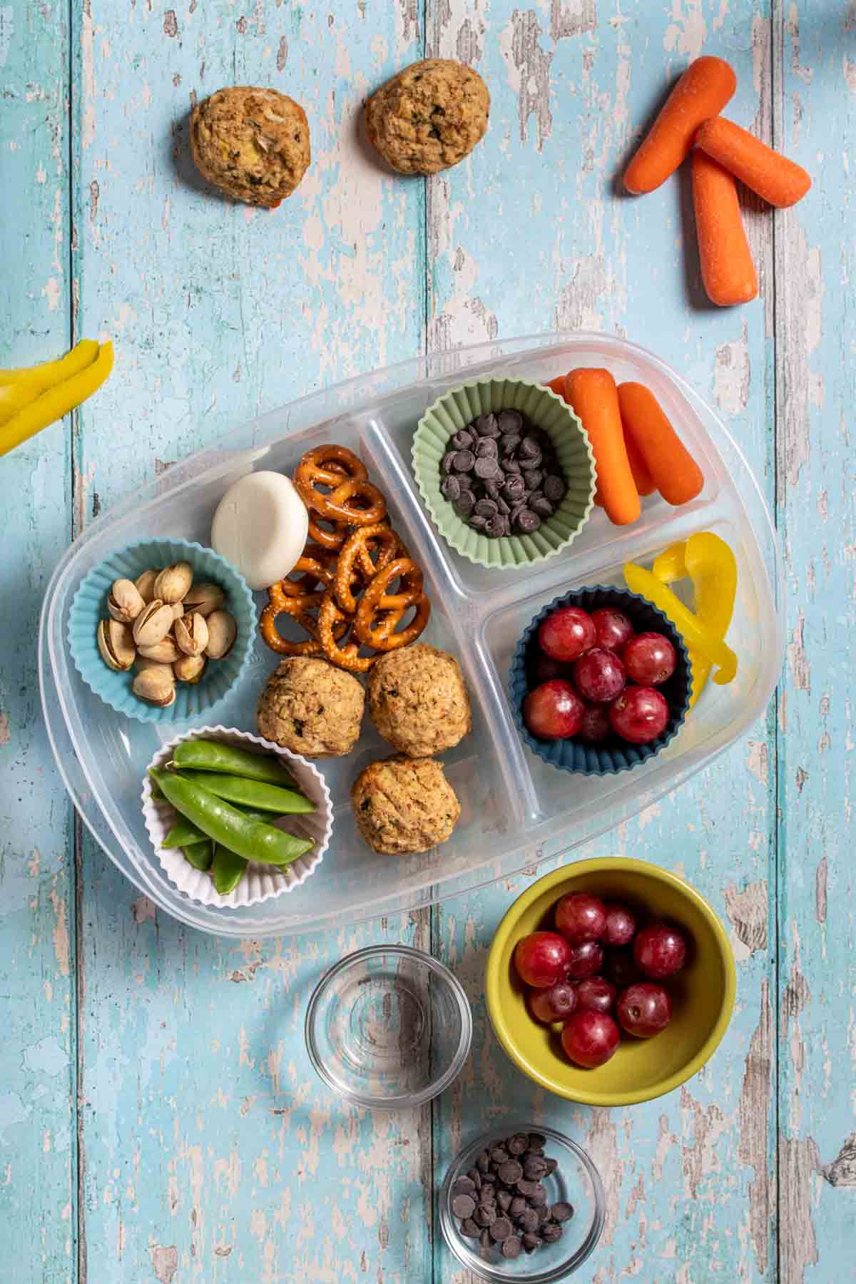 Top view of a container with sections filled with veggies, fruit, pretzels, nuts and protein balls on a light blue surface.