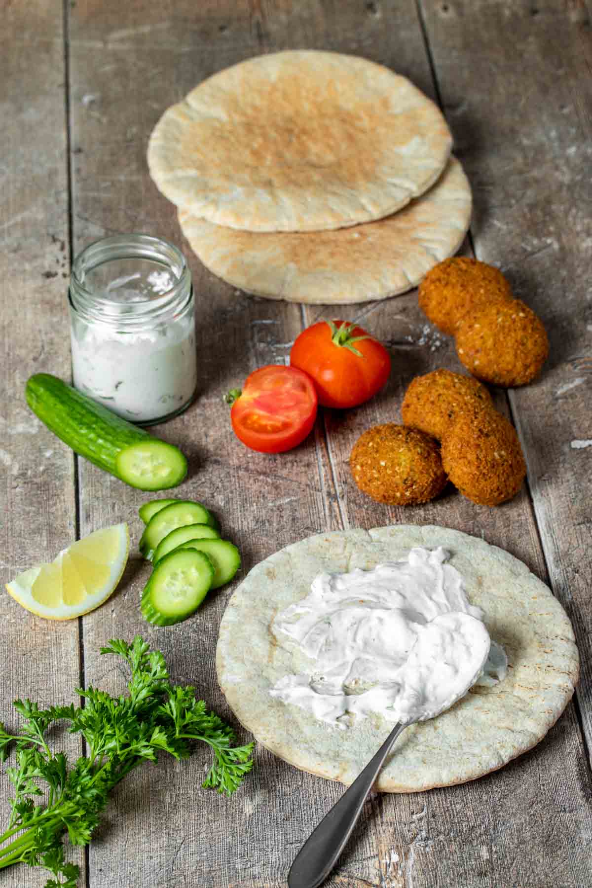 A wooden table with ingredients to make a falafel pita and one pita with sauce on it next to them.