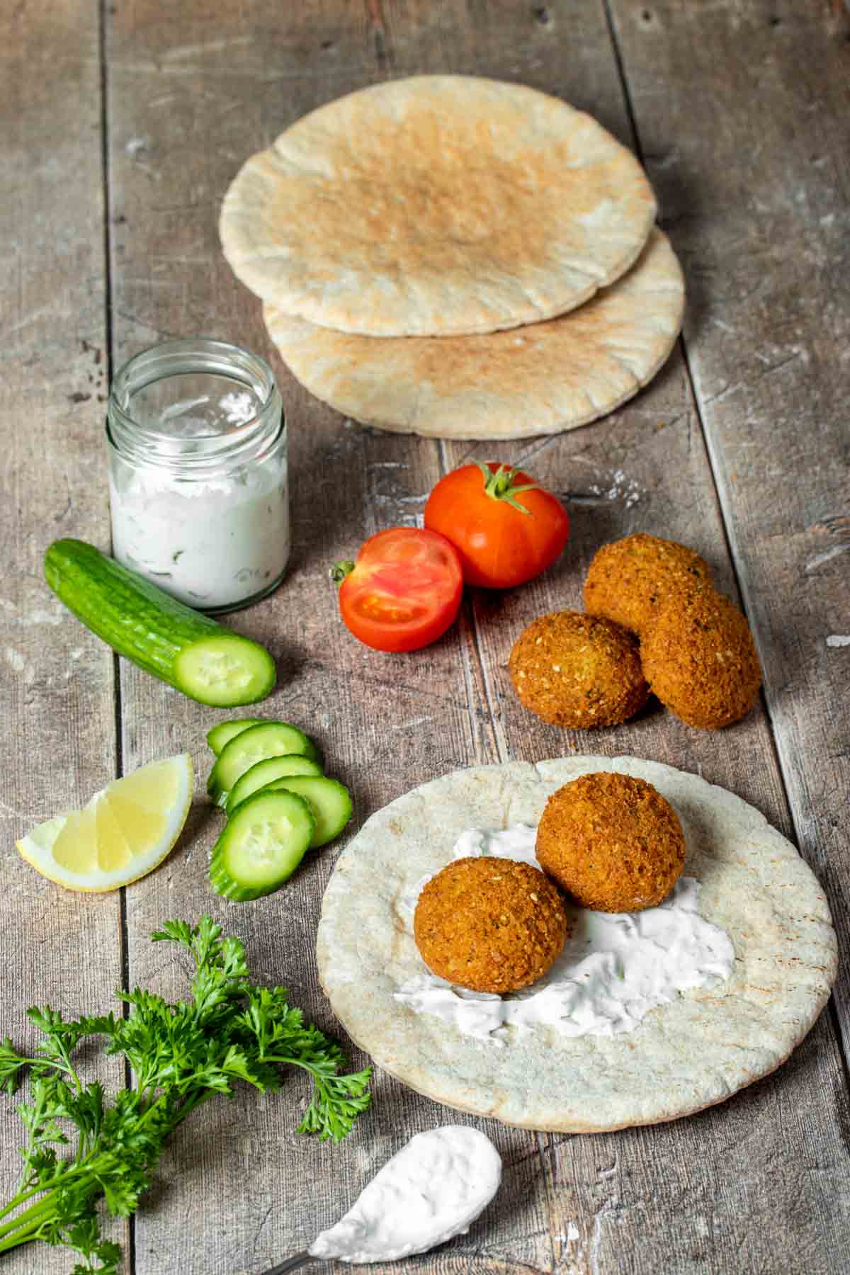 Pita bread spread with sauce and two falafel with more ingredients around it on a wooden table.