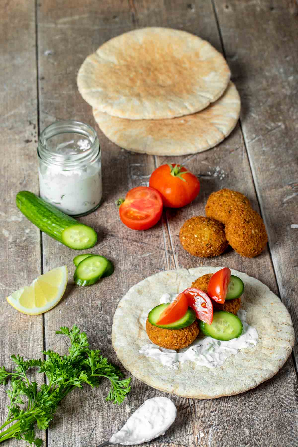 A pita bread with sauce, falafel, cucumbers and tomatoes on it surrounded by more of the imgredients.
