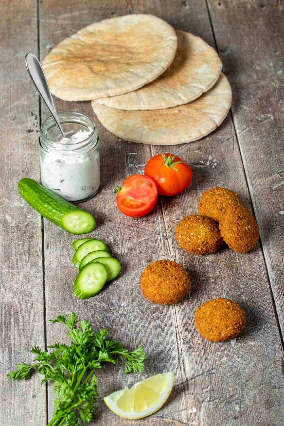 A wooden table with pita bread, tzatziki sauce in a jar, cucumber, tomato, falafel, parsley and a lemon wedge.