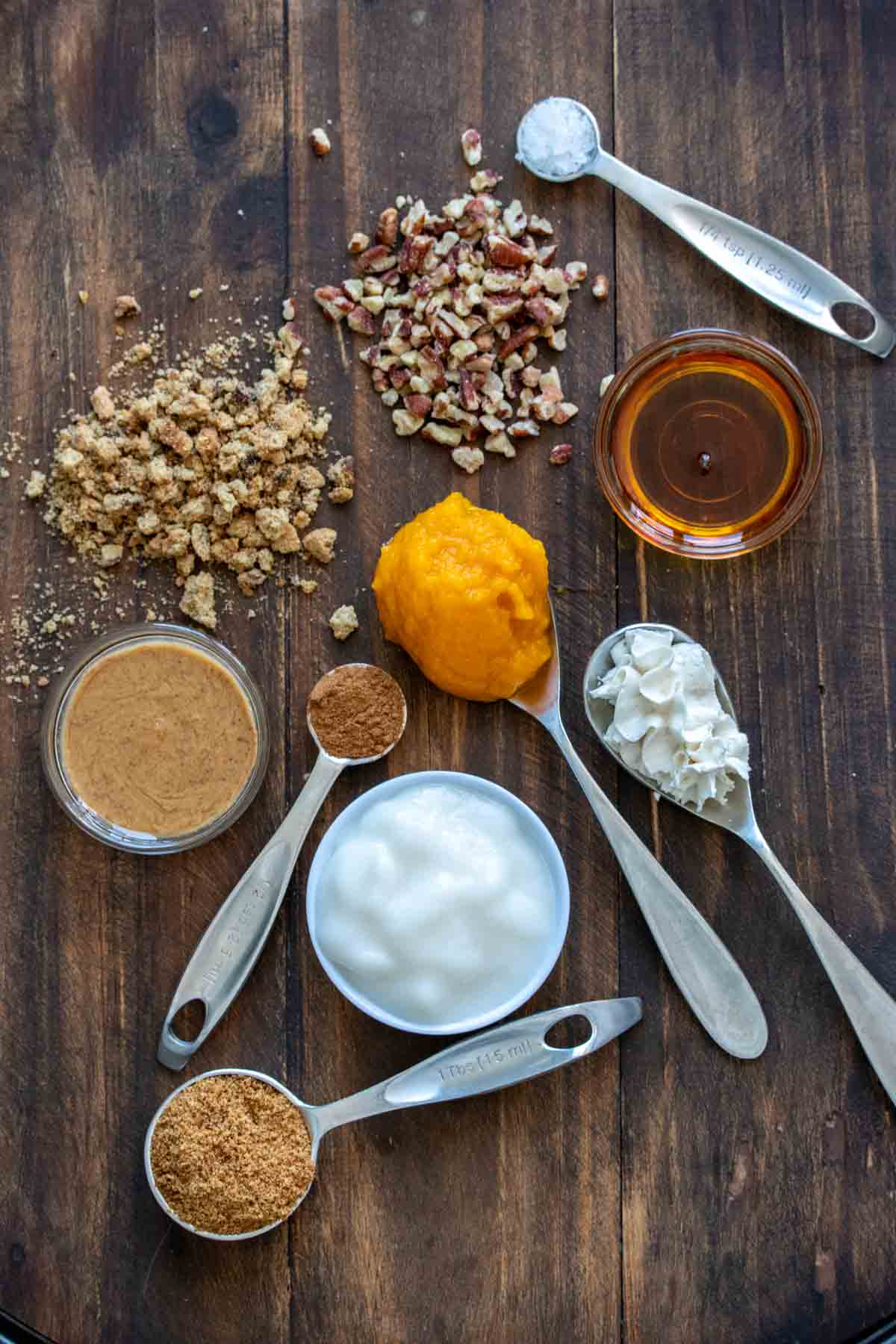 Spices, yogurt, pumpkin, whipped cream, nuts and crumbled cookies in spoons, bowls and on a wooden surface.