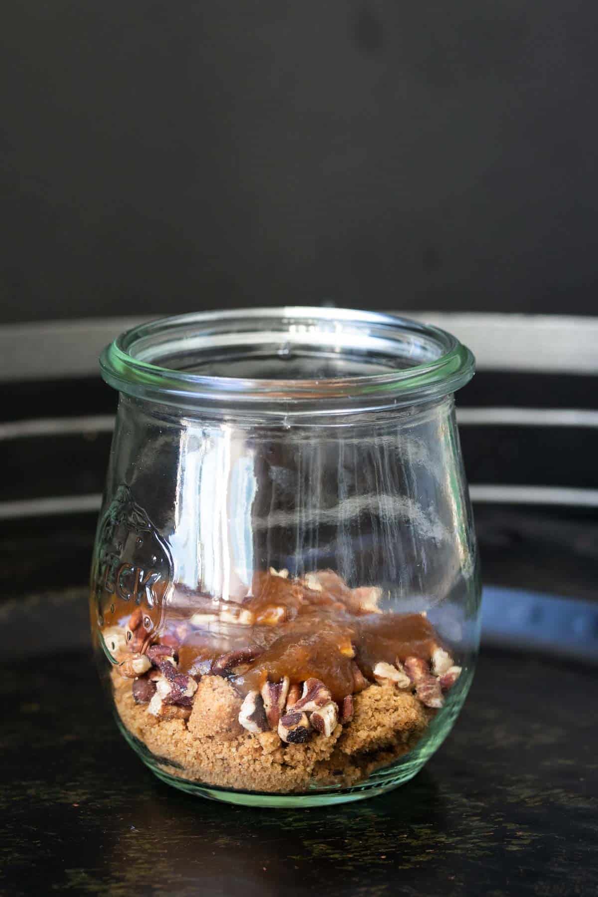 A glass jar layered with crushed cookies, pecans and caramel.