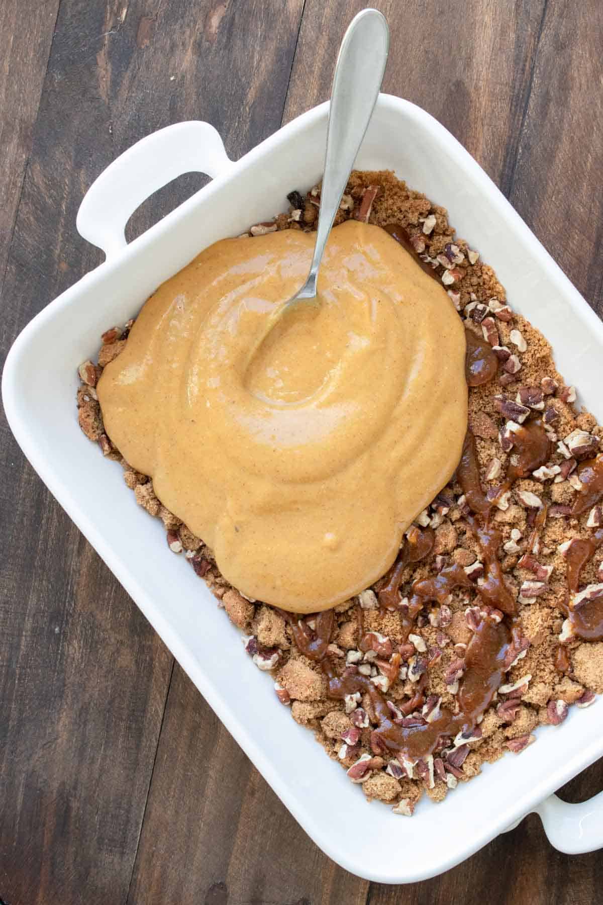 A creamy pumpkin filling being spread over nuts and cookie pieces in a white baking dish.