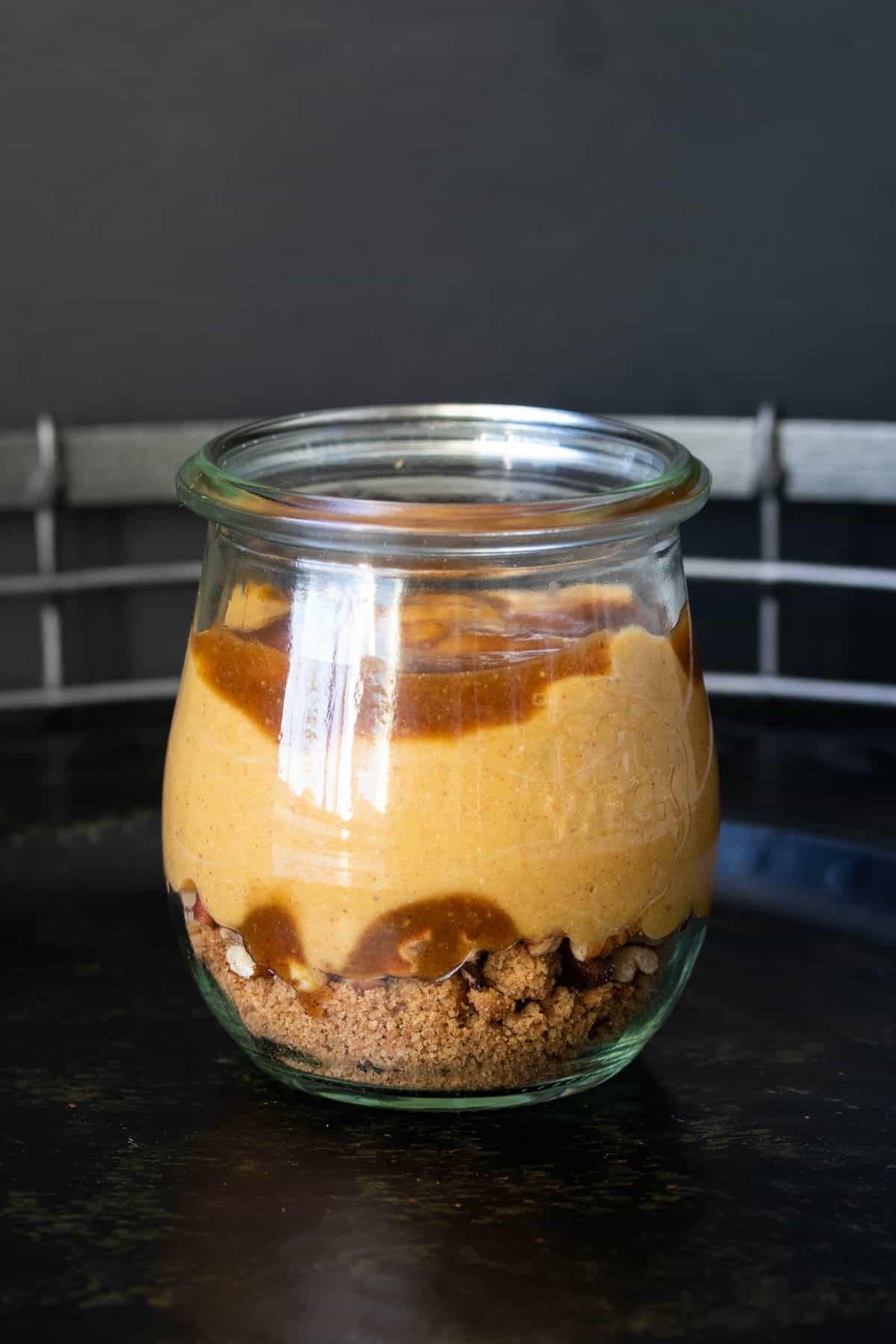 A glass jar layered with crushed cookies, nuts, caramel and a creamy pumpkin filling.
