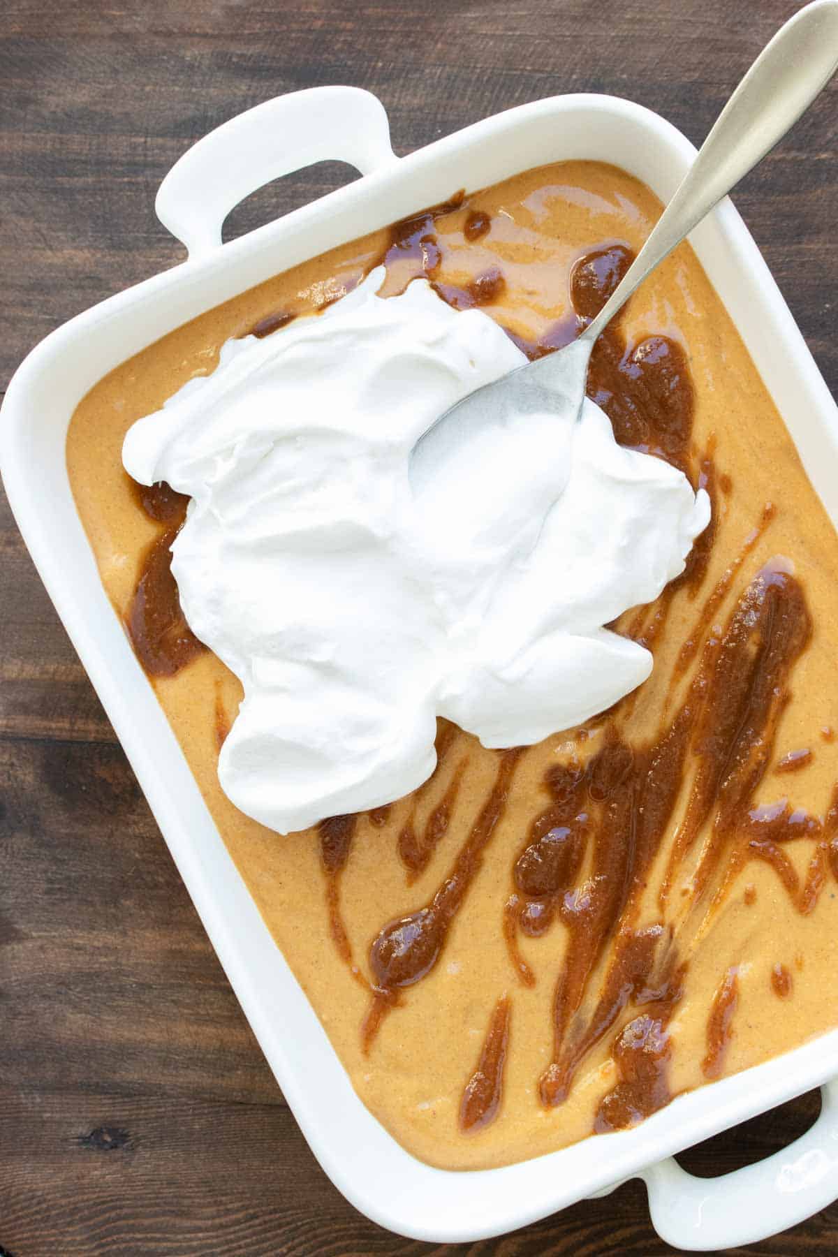 A spoon spreading whipped cream over a creamy pumpkin filling drizzled with caramel.