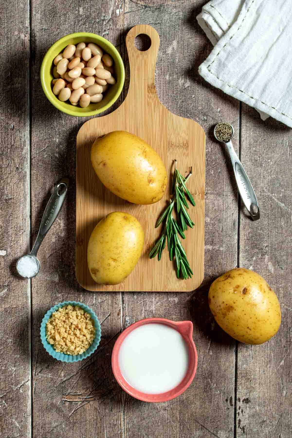 Top view of a wooden surface with potatoes and rosemary on a cutting board surrounded by beans, milk, parmesan and spices.