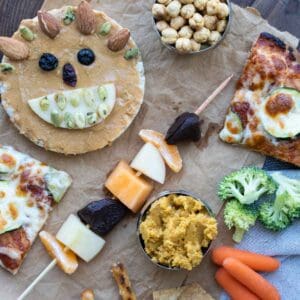 A rice cake decorated with food to make a face next to a fruit skewer, veggies, hummus, pizza and chickpeas on a piece of parchment paper.