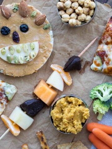 A rice cake decorated with food to make a face next to a fruit skewer, veggies, hummus, pizza and chickpeas on a piece of parchment paper.