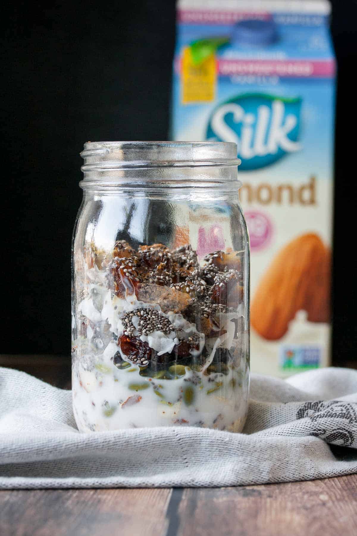 A jar with dried fruits, seeds, nuts and oats with milk at the bottom in front of a colorful container of almond milk.