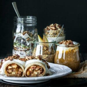 A white plate with apple crepes, jars of pumpkin and pear flavored oats and pudding and a jar with cereal inside.