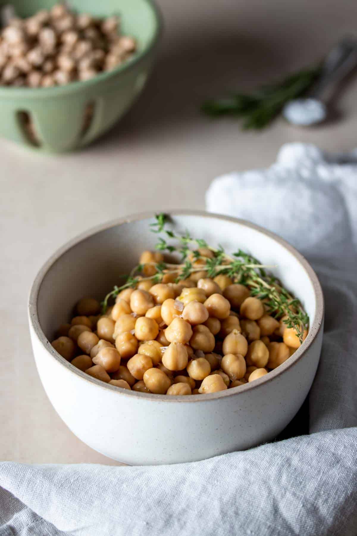 Cooked chickpeas in a white bowl with tan rim and sprigs of thyme with a green strainer in the background.