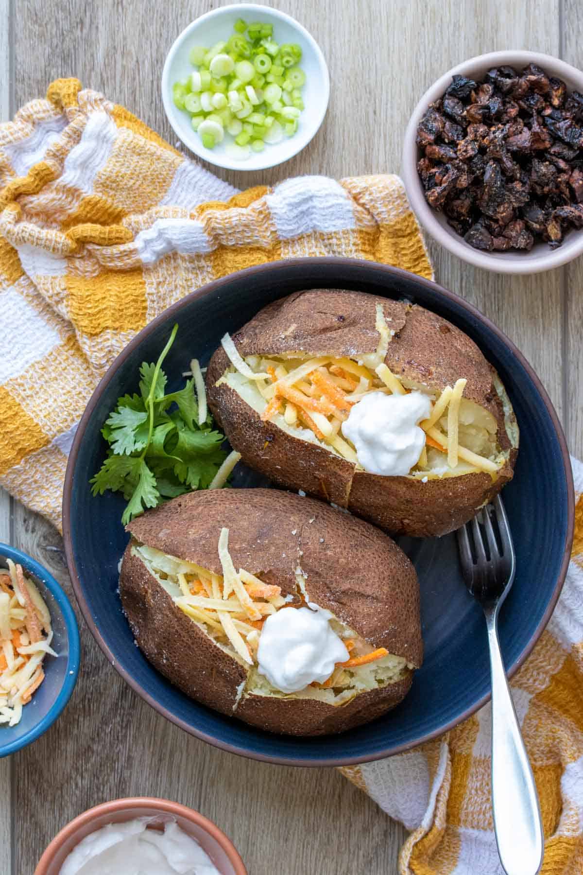 Shredded cheese and sour cream on two baked potatoes in a blue shallow bowl surrounded by other ingredients.