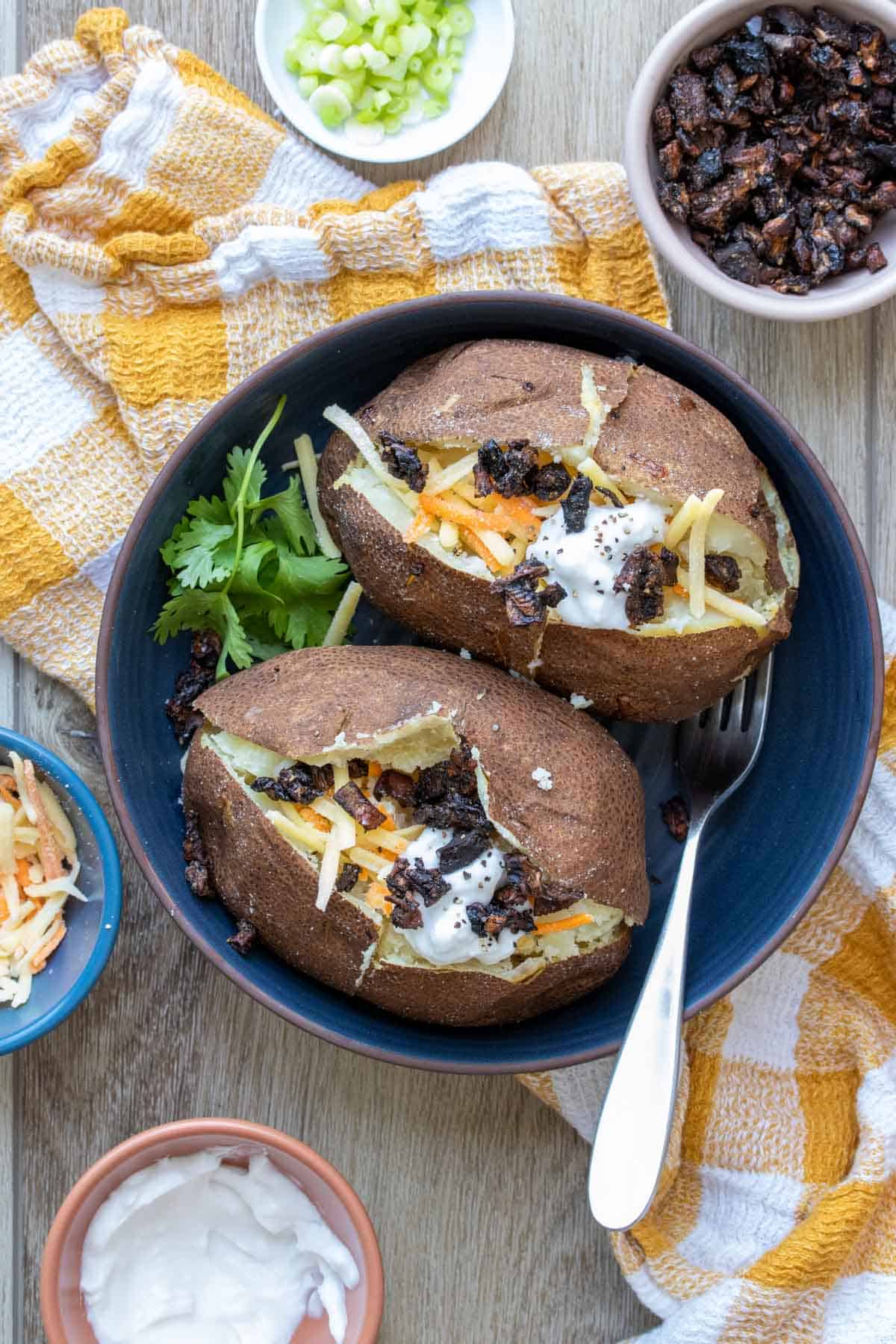 Two baked potatoes in a shallow blue bowl topped with cheese, sour cream and chopped portobello bacon on a yellow checkered towel.