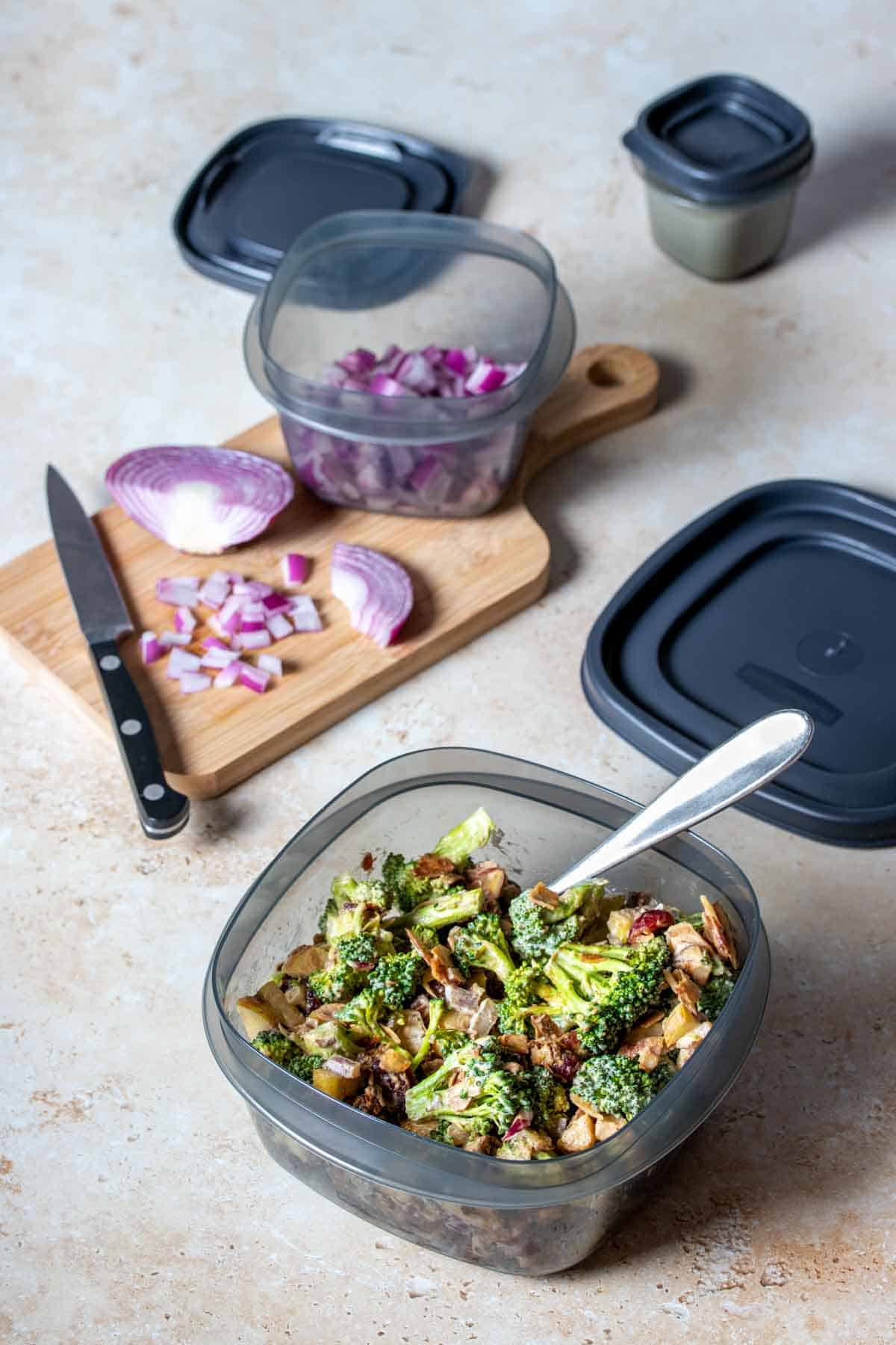 A grey plastic container filled with broccoli salad on a tan surface in front of a smaller container with chopped red onion.