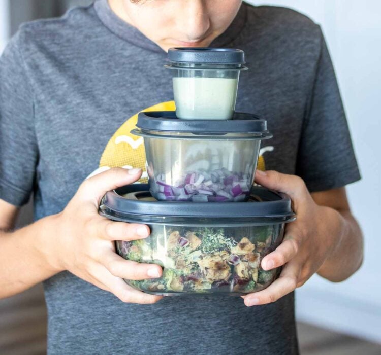 A boy holding three different sized grey plastic containers with things in them looking down at the stack in his hands.