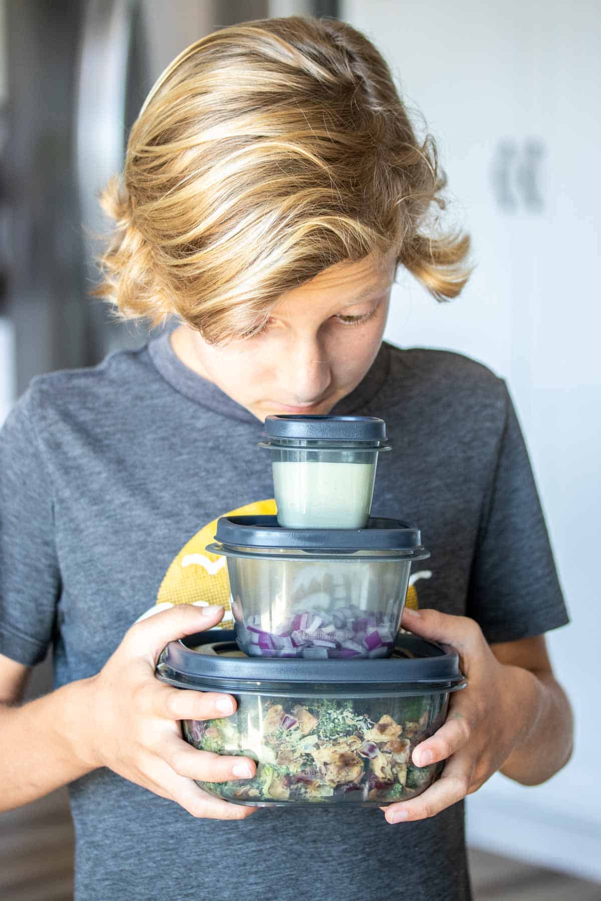 A boy holding three different sized grey plastic containers with things in them looking down at the stack in his hands.