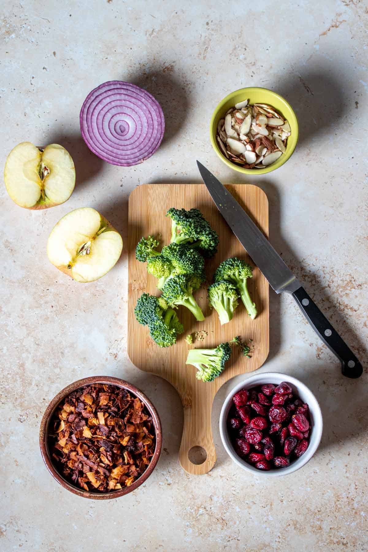 A small wooden cutting board with cut broccoli next to apples, onion, slivered almonds, dried cranberries and coconut bacon.