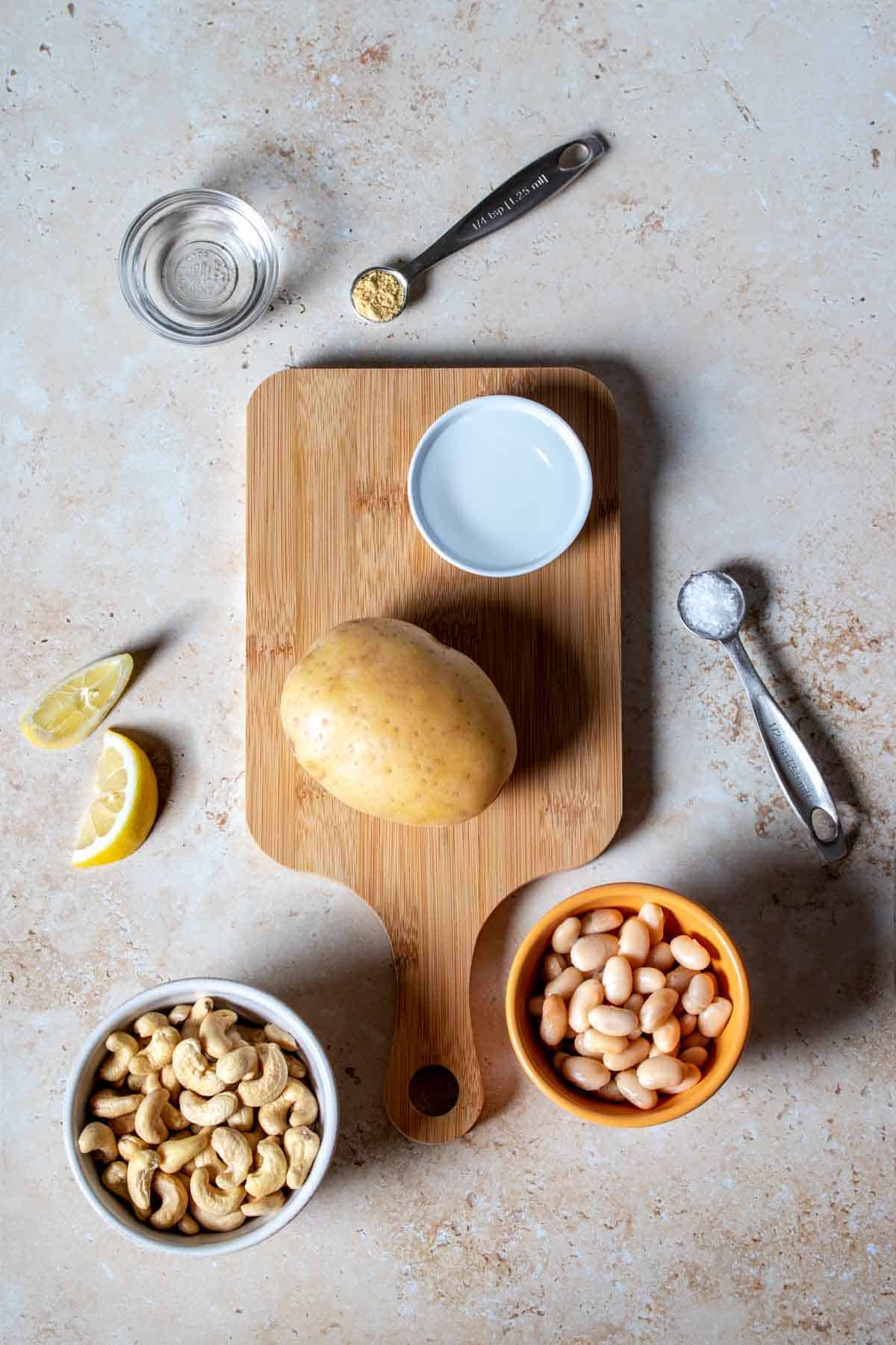 Bowls of cashews and white beans next to a small cutting board with a potato and small bowl of clear liquid and pieces of lemon, spices and more clear liquid in a small glass bowl on the surface.