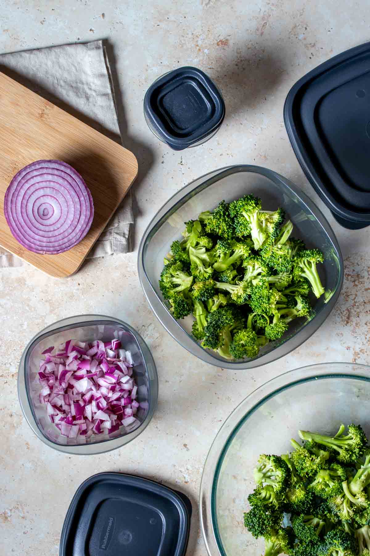 Top view of cut broccoli and onion in plastic containers next to their lids on a tan surface.