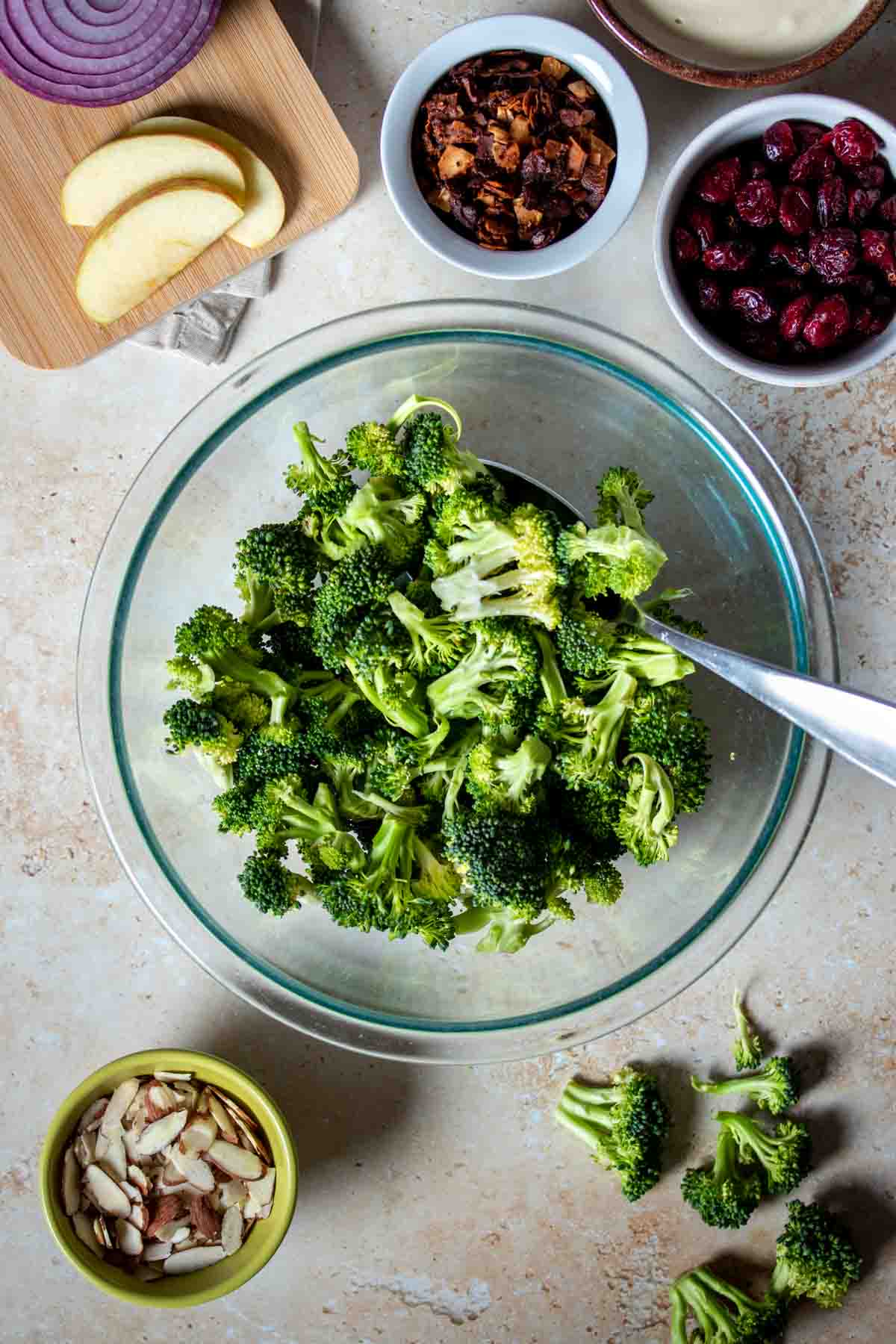 A glass bowl with chopped broccoli next to ingredients to make a classic broccoli salad.