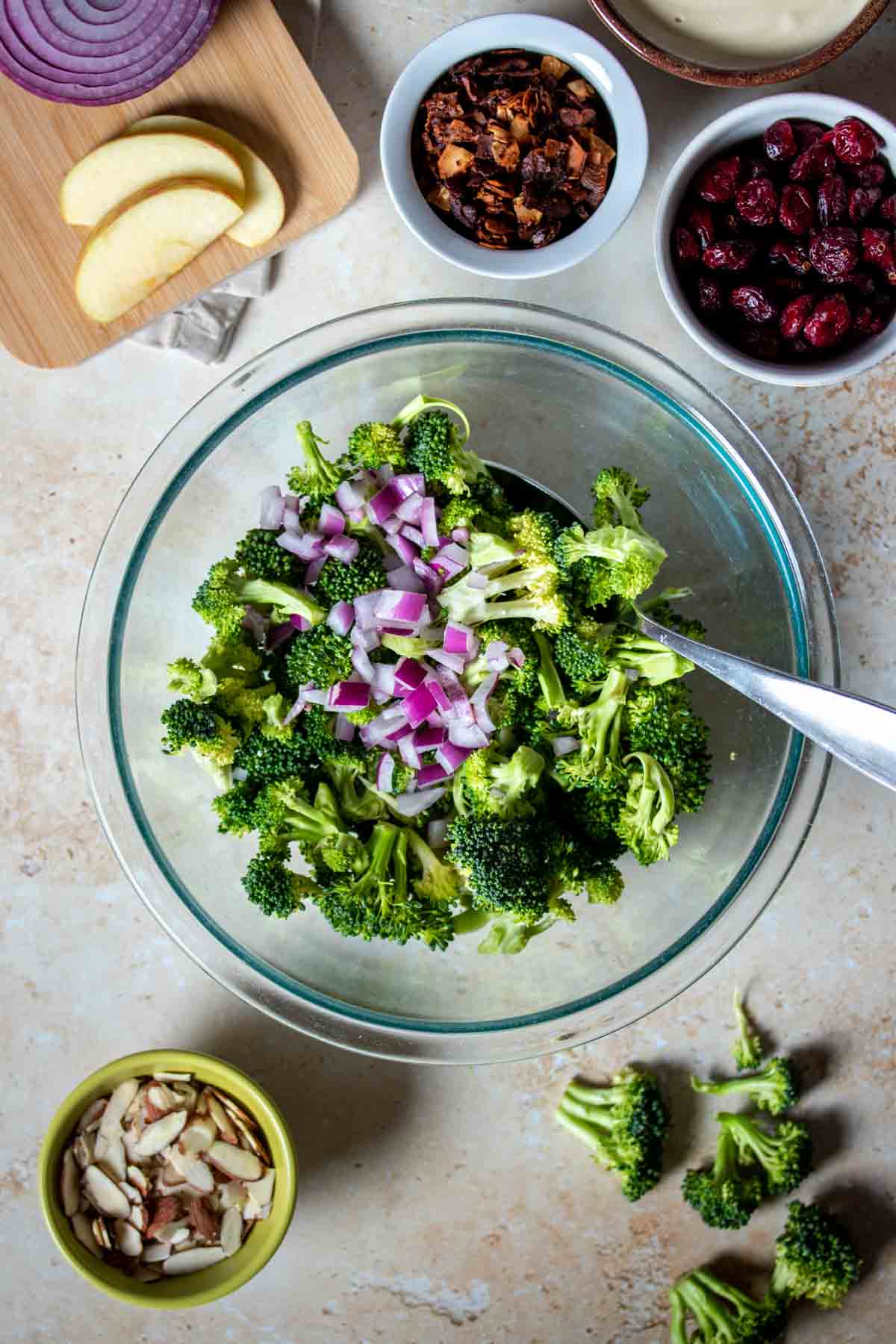 Chopped red onion and broccoli in a glass bowl next to other ingredients for a broccoli salad.