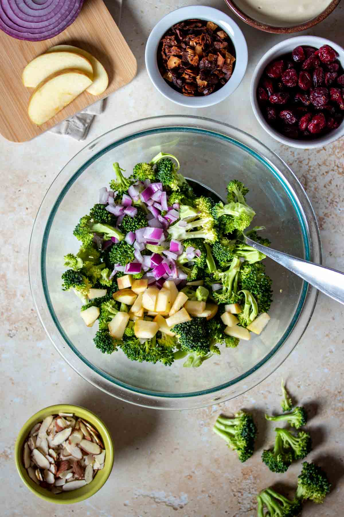 Chopped apples, red onion and broccoli in a glass bowl next to bowls of ingredients for a broccoli salad.