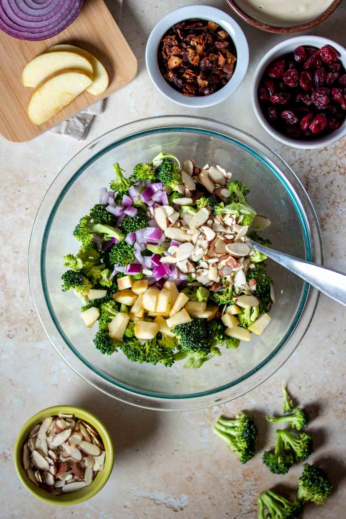 Chopped apples, red onion, broccoli and slivered almonds in a glass bowl next to more of the ingredients for the broccoli salad.