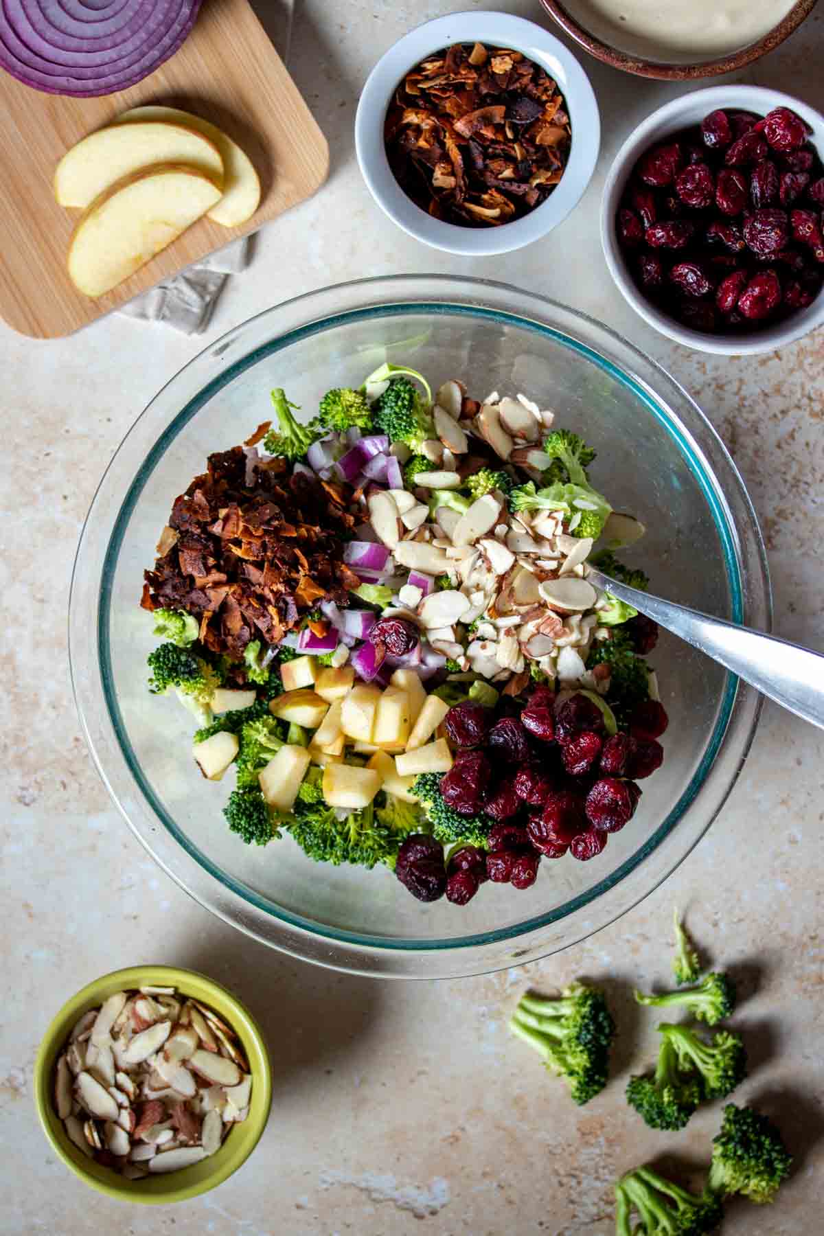 Coconut bacon, cranberries, almonds, red onion, apples and broccoli in a glass bowl surrounded by more of them in bowls on a tan surface.