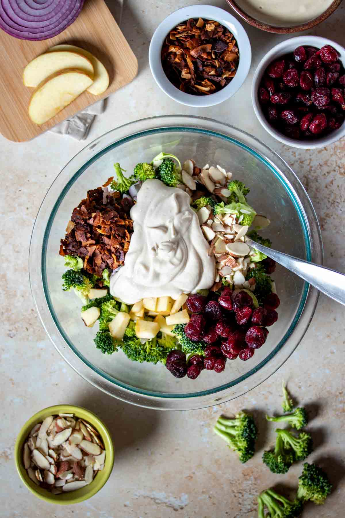 A creamy dressing over ingredients for a broccoli salad in a large glass bowl with more ingredients around the bowl.