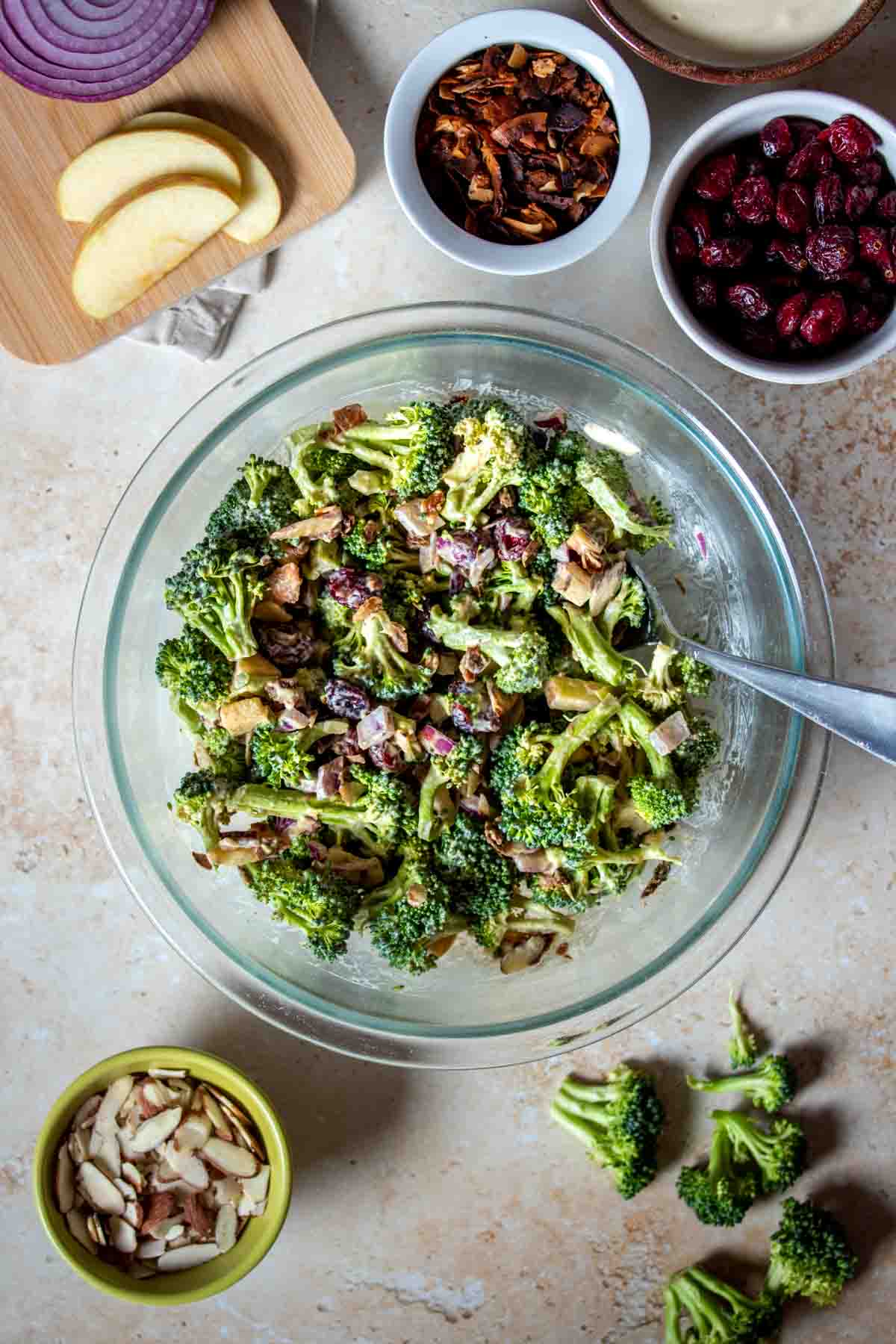 A creamy broccoli salad in a glass bowl being mixed by a spoon surrounded by more ingredients in smaller bowls.