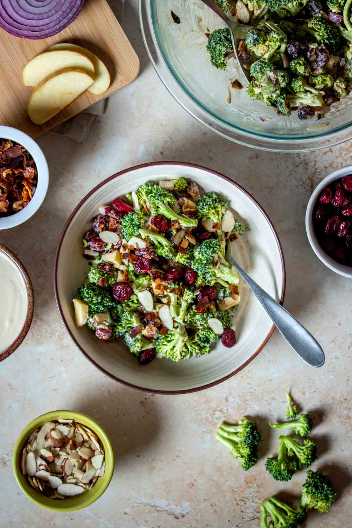 A creamy broccoli salad with cranberries in a tan bowl with a brown rim surrounded by ingredients and more salad.