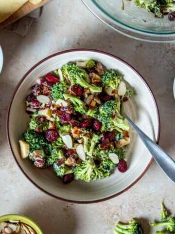 A creamy broccoli salad topped with cranberries and slivered almonds in a tan bowl with a fork.