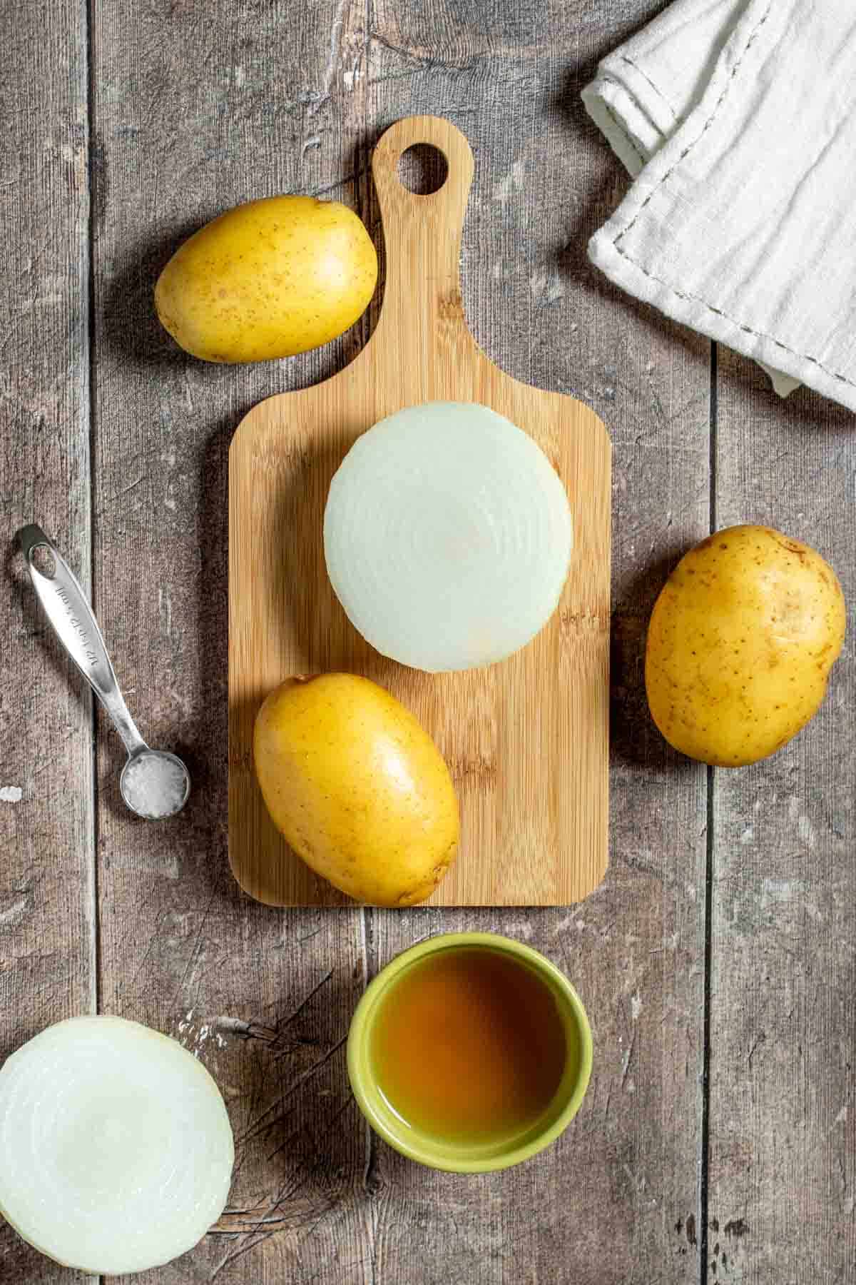 Top view of a wooden surface with a cutting board, potatoes, onion, salt and broth in a bowl.